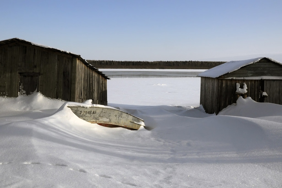 Ein kleines Ruderboot liegt teilweise im tiefen Schnee zwischen zwei hölzernen Bootsschuppen.